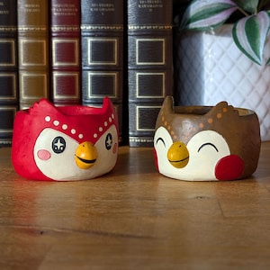May include: Two owl-shaped ceramic planters. One is red with white accents, a yellow beak, and a smiling face. The other is brown with white, red, and yellow accents. Both have a cartoonish design and are suitable for small plants.