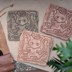 以下が含まれることがあります： 漫画のキャラクターデザインが施された、正方形のテクスチャードストーンカービングが3つ。彫刻は茶色とグレーの色合いです。彫刻は、アートツールと緑のあるニュートラルな表面に配置されています。