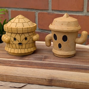 May include: Two tan ceramic figurines with cartoon faces and arms. One has a domed lid, the other a conical top. They sit on a wooden board against a brick background. The handcrafted figures have a unique, whimsical design.