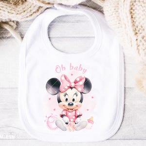 K&ouml;nnte beinhalten: Wei&szlig;es Baby-L&auml;tzchen mit Minnie Mouse-Design. Das L&auml;tzchen hat eine rosa Schleife mit Punkten, ein rosa Outfit und die Worte "Oh baby" in rosa Schrift. Eine Babyflasche und Bl&ouml;cke sind ebenfalls im Design enthalten.