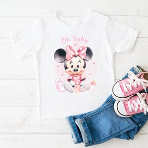 K&ouml;nnte beinhalten: Wei&szlig;es T-Shirt mit Minnie Mouse-Grafik und dem Text "Oh baby". Das Design umfasst eine rosa Schleife, ein Kleid und Details. Dazu blaue Jeans und rosa Turnschuhe.