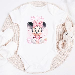 K&ouml;nnte beinhalten: Wei&szlig;er Baby-Body mit einem Minnie Mouse-Motiv in Rosa und Schwarz. Der Body tr&auml;gt den rosa Schriftzug "Oh baby". Wei&szlig;e Babyschuhe und ein wei&szlig;es H&auml;schen-Rassel aus H&auml;kelarbeit sind ebenfalls im Bild.