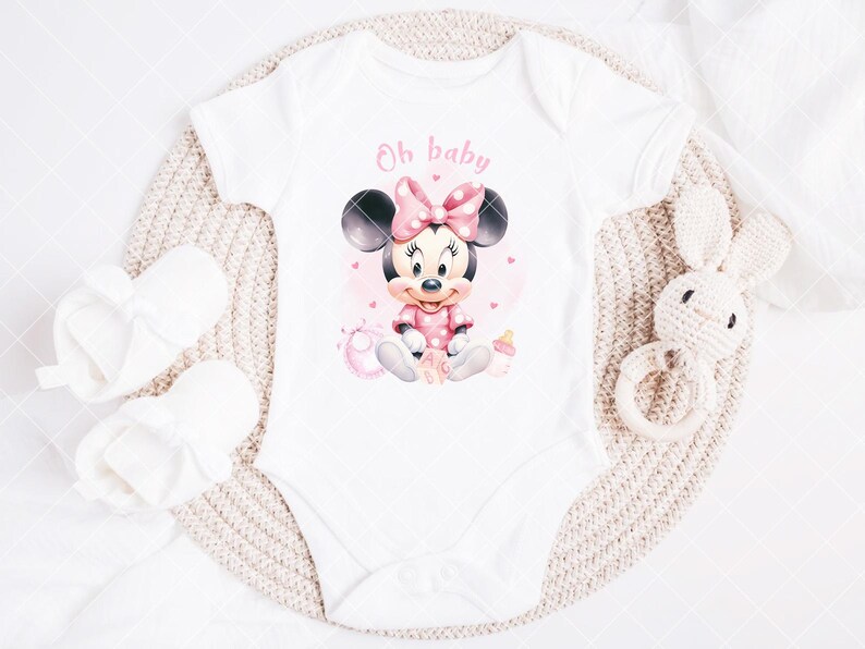 K&ouml;nnte beinhalten: Wei&szlig;er Baby-Body mit einem Minnie Mouse-Motiv in Rosa und Schwarz. Der Body tr&auml;gt den rosa Schriftzug "Oh baby". Wei&szlig;e Babyschuhe und ein wei&szlig;es H&auml;schen-Rassel aus H&auml;kelarbeit sind ebenfalls im Bild.