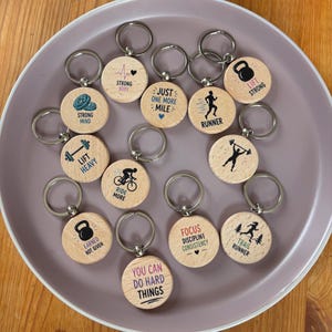 Könnte beinhalten: Eine Sammlung runder Holzschlüsselanhänger mit verschiedenen Fitness-Grafiken und motivierenden Texten wie Strong Body, Just One More Mile, Runner, Lift Strong, Strong Mind, Lift Heavy, Ride More, Earned Not Given, Focus Discipline Consistency, Trail Runner und You Can Do Hard Things.