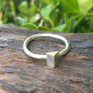 Peut inclure: Une bague en argent avec un motif géométrique de forme carrée. La bague est posée sur une surface en bois.