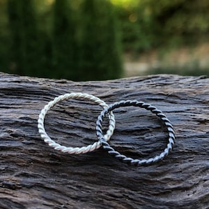May include: Two thin, twisted rings, one silver and one black, are lying on a piece of weathered wood.