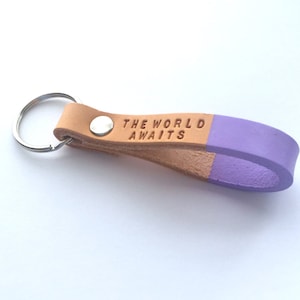 Puede incluir: Un llavero de cuero marrón con un lazo de cuero morado. El llavero tiene el texto "THE WORLD AWAITS" grabado en relieve.