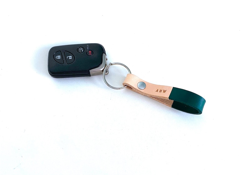 May include: A tan leather keychain with a green leather strap and a silver key ring. The strap has the initials "AMW" embossed on it.
