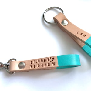 May include: Two leather keychains with a light brown strap and a turquoise blue accent. One keychain has a silver key ring and the other has a silver clasp. The keychain with the clasp has the coordinates "38.89997&deg;N 77.0486&deg;W" engraved on the strap. The other keychain has the initials "L.F.H." engraved on the strap.