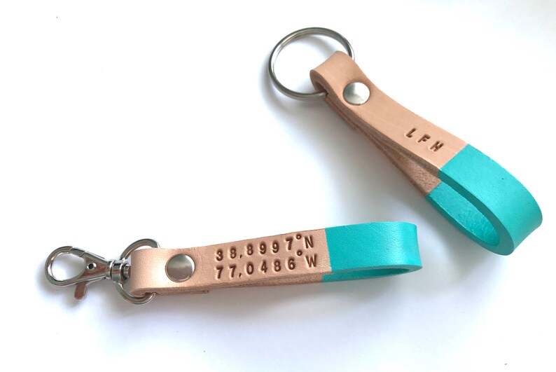 May include: Two leather keychains with a light brown strap and a turquoise blue accent. One keychain has a silver key ring and the other has a silver clasp. The keychain with the clasp has the coordinates "38.89997&deg;N 77.0486&deg;W" engraved on the strap. The other keychain has the initials "L.F.H." engraved on the strap.