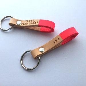 Puede incluir: Dos llaveros de cuero con detalles de color rosa brillante. Un llavero tiene el texto "YOU GO, GLEN COCO!" y el otro las iniciales "JMG".