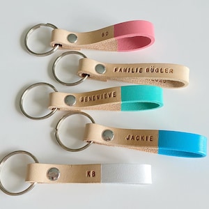 May include: Five leather keychains with different colored tips. The keychains are beige with a silver key ring. The tips are pink, turquoise, blue, white, and a beige with the letters "KB" embossed on it. The keychains have the following names embossed on them: "BD", "FAMILIE B&Uuml;GLER", "GENEVIEVE", "JACKIE", and "KB".
