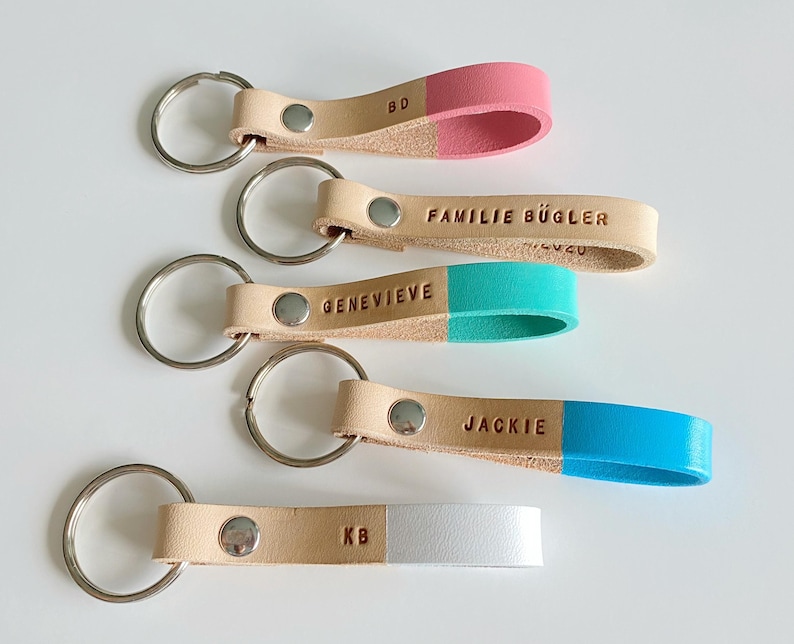 May include: Five leather keychains with different colored tips. The keychains are beige with a silver key ring. The tips are pink, turquoise, blue, white, and a beige with the letters "KB" embossed on it. The keychains have the following names embossed on them: "BD", "FAMILIE B&Uuml;GLER", "GENEVIEVE", "JACKIE", and "KB".