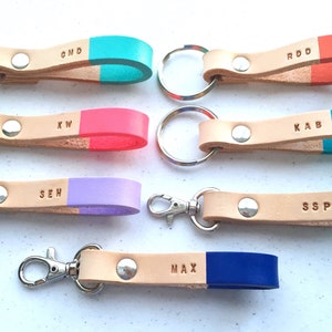 May include: Eight leather keychains with different colored tips and personalized initials. The keychains are made of natural leather and have a silver key ring or hook.