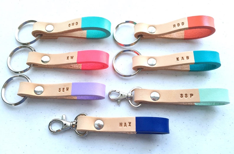 May include: Eight leather keychains with different colored tips and personalized initials. The keychains are made of natural leather and have a silver key ring or hook.