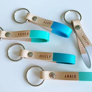 May include: Five leather keychains with personalized names, Kory, Rhonda, Cecily, Abbie, and Ashley. Each keychain has a light brown leather strap with a contrasting color on the bottom half, either turquoise or white.