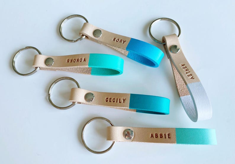 May include: Five leather keychains with personalized names, Kory, Rhonda, Cecily, Abbie, and Ashley. Each keychain has a light brown leather strap with a contrasting color on the bottom half, either turquoise or white.