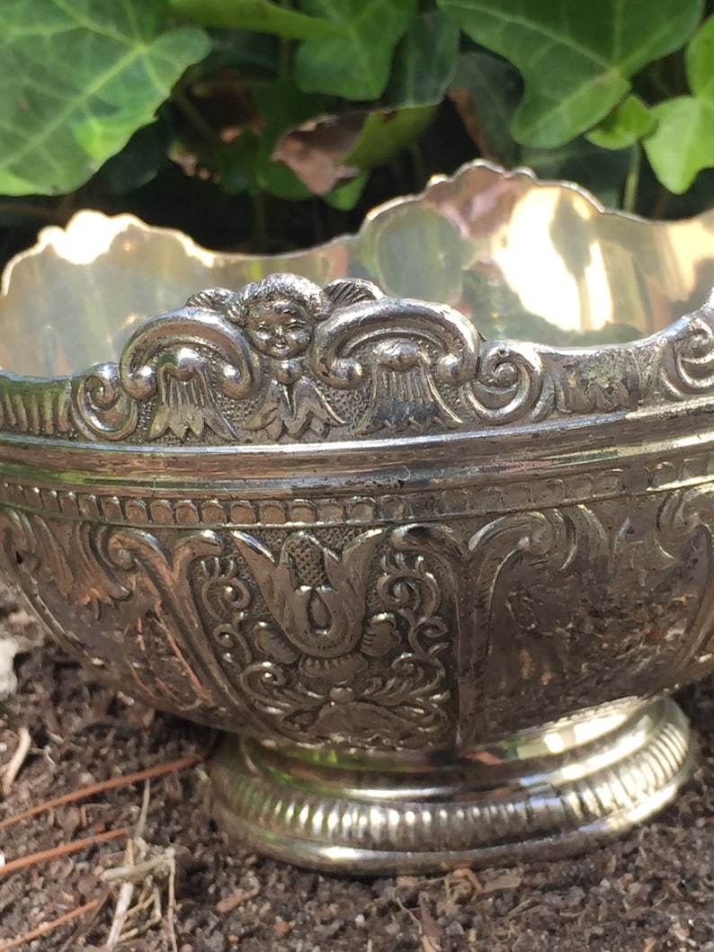 May include: A silver bowl with an ornate design featuring cherubs and floral motifs. The bowl has a wide rim and a round base.
