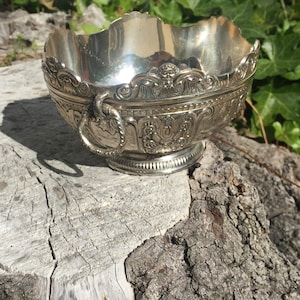 May include: A silver bowl with an ornate design and a handle. The bowl is sitting on a tree stump.