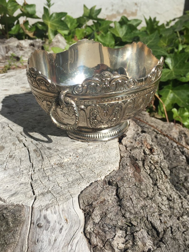 May include: A silver bowl with an ornate design and a handle. The bowl is sitting on a tree stump.