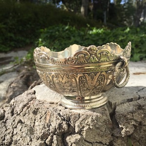 May include: A silver bowl with intricate floral and scrollwork designs. The bowl has a handle on one side and sits on a wooden stump.