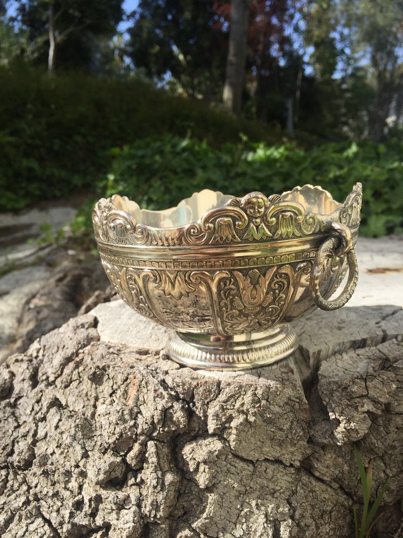 May include: A silver bowl with intricate floral and scrollwork designs. The bowl has a handle on one side and sits on a wooden stump.