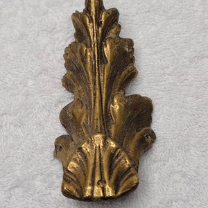 May include: A gold-colored metal furniture leg with a decorative, ornate, leaf-like design.