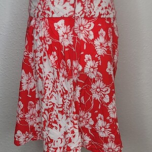 May include: A red and white floral print skirt. The skirt features a bold red background with a repeating pattern of white flowers. The waistband is white with a matching floral design. The skirt is displayed on a mannequin.