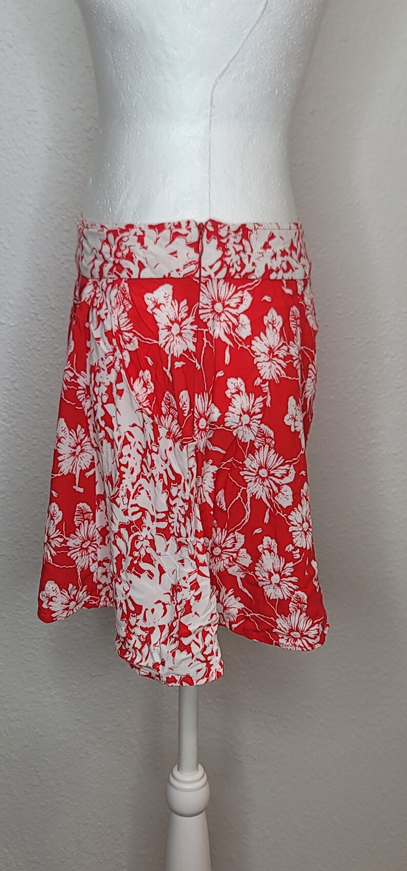 May include: A red and white floral print skirt. The skirt features a bold red background with a repeating pattern of white flowers. The waistband is white with a matching floral design. The skirt is displayed on a mannequin.