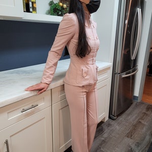 Peut inclure: Un tailleur rose clair, composé d'une veste ajustée avec plusieurs boutons et poches, et d'un pantalon large assorti. Le costume a un aspect sur mesure avec des coutures visibles. La personne porte des chaussures noires.