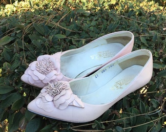 May include: A pair of pink leather heeled shoes with a flower design on the toe. The shoes have a white sole and are sitting on a bed of green leaves.