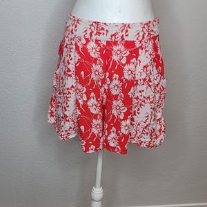 May include: Red and white floral print shorts displayed on a white mannequin. The shorts feature a vibrant red background with a repeating pattern of white flowers. The garment is made of a lightweight fabric, perfect for warm weather.