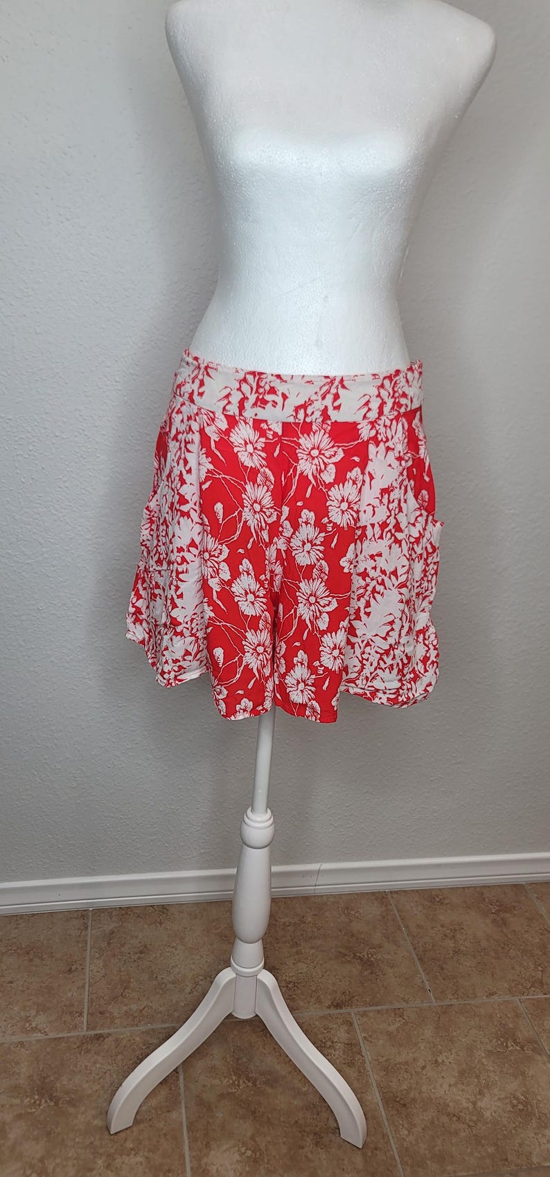 May include: Red and white floral print shorts displayed on a white mannequin. The shorts feature a vibrant red background with a repeating pattern of white flowers. The garment is made of a lightweight fabric, perfect for warm weather.