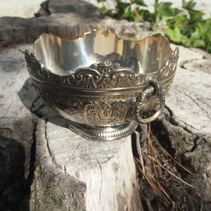 May include: A silver bowl with an ornate design and a handle. The bowl is sitting on a piece of wood.