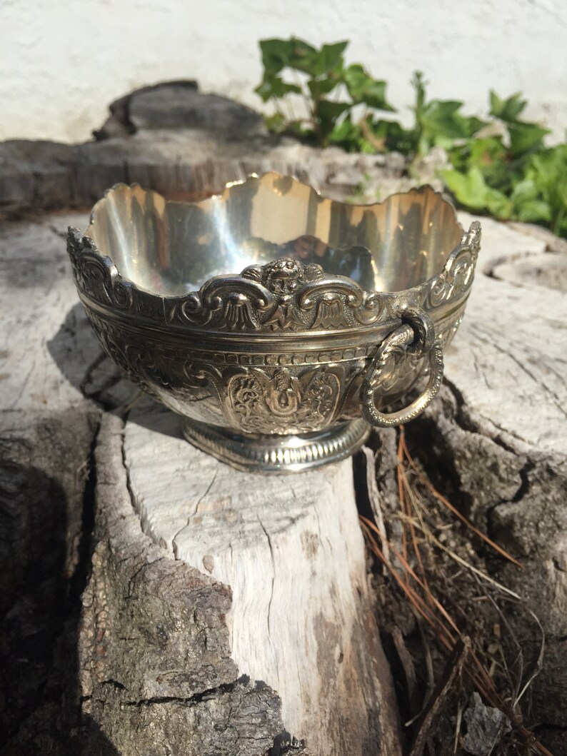 May include: A silver bowl with an ornate design and a handle. The bowl is sitting on a piece of wood.