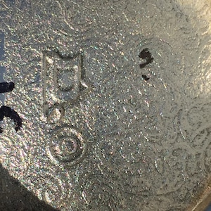 May include: Close-up of a silver-coloured metal object with a circular mark and a rectangular mark containing the letter "D". The object appears to be a vintage or antique item.