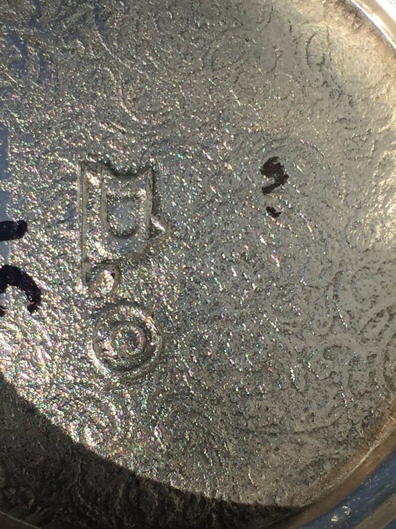 May include: Close-up of a silver-coloured metal object with a circular mark and a rectangular mark containing the letter "D". The object appears to be a vintage or antique item.