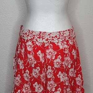 May include: Red shorts with a white floral pattern. The shorts are displayed on a white mannequin. The shorts have a white waistband and a ruffled hem. The shorts are made of a lightweight fabric.