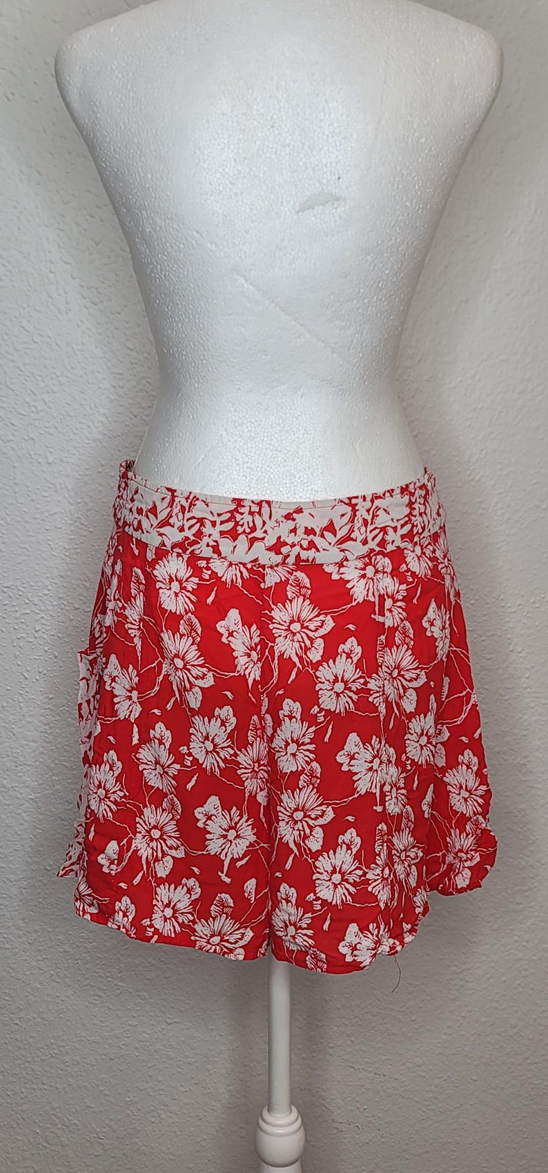 May include: Red shorts with a white floral pattern. The shorts are displayed on a white mannequin. The shorts have a white waistband and a ruffled hem. The shorts are made of a lightweight fabric.