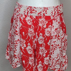 May include: Red and white floral print shorts. The shorts feature a vibrant red background with a white floral pattern. The shorts are displayed on a mannequin, showcasing their design and fit. The shorts have pockets.