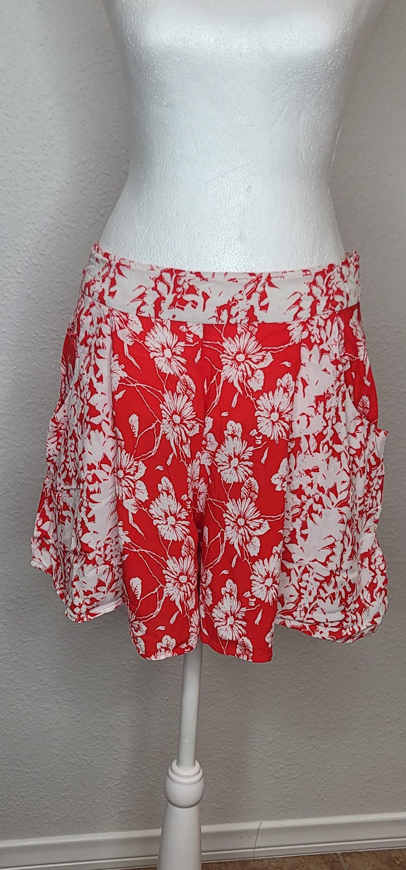 May include: Red and white floral print shorts. The shorts feature a vibrant red background with a white floral pattern. The shorts are displayed on a mannequin, showcasing their design and fit. The shorts have pockets.