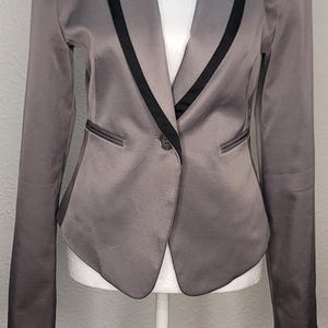 Puede incluir: Un blazer gris con ribete negro sobre un maniquí blanco. El blazer tiene un cierre de un solo botón y dos bolsillos delanteros. Las solapas y el cuello están delineados con un fino ribete negro. La tela tiene un ligero brillo.