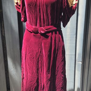 May include: A deep burgundy velvet dress featuring short puffed sleeves with cream-colored floral embroidery on the shoulders, a gathered neckline, and a matching velvet belt with a central knot detail at the waist, falling to a midi length.