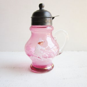 May include: A pink glass creamer with a silver lid and a painted design of a person riding a bicycle. The creamer has a gold trim around the base.