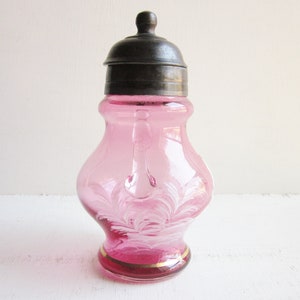 May include: A pink glass sugar dispenser with a silver lid and a white floral design. The dispenser has a small handle on the side.