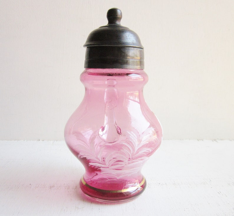 May include: A pink glass sugar dispenser with a silver lid and a white floral design. The dispenser has a small handle on the side.