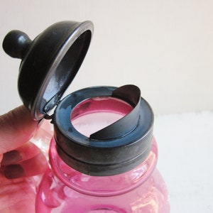 May include: A pink glass bottle with a silver lid. The lid is hinged and has a small spout. The bottle is likely for storing liquids.