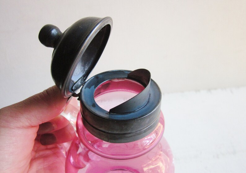 May include: A pink glass bottle with a silver lid. The lid is hinged and has a small spout. The bottle is likely for storing liquids.