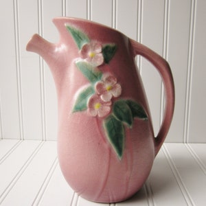 May include: A pink ceramic pitcher with a floral design. The pitcher has a spout and a handle. The flowers are pink and green.