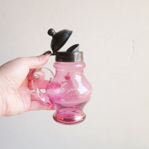 May include: A pink glass creamer with a silver lid and a bicycle design. The creamer has a gold rim.